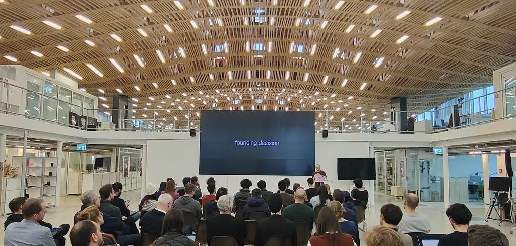 An audience under a timber roof listening to a talk, a screen reading “founding decision”