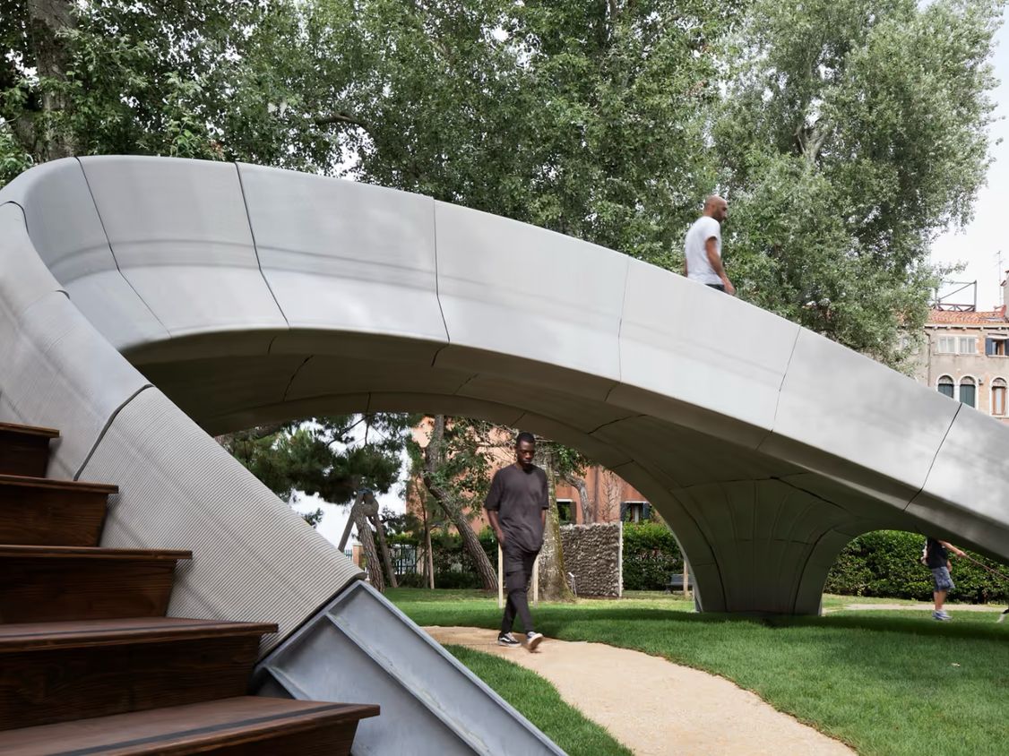 A grey arched concrete footbridge with timber steps in a tree-lined garden, people walking beneath it