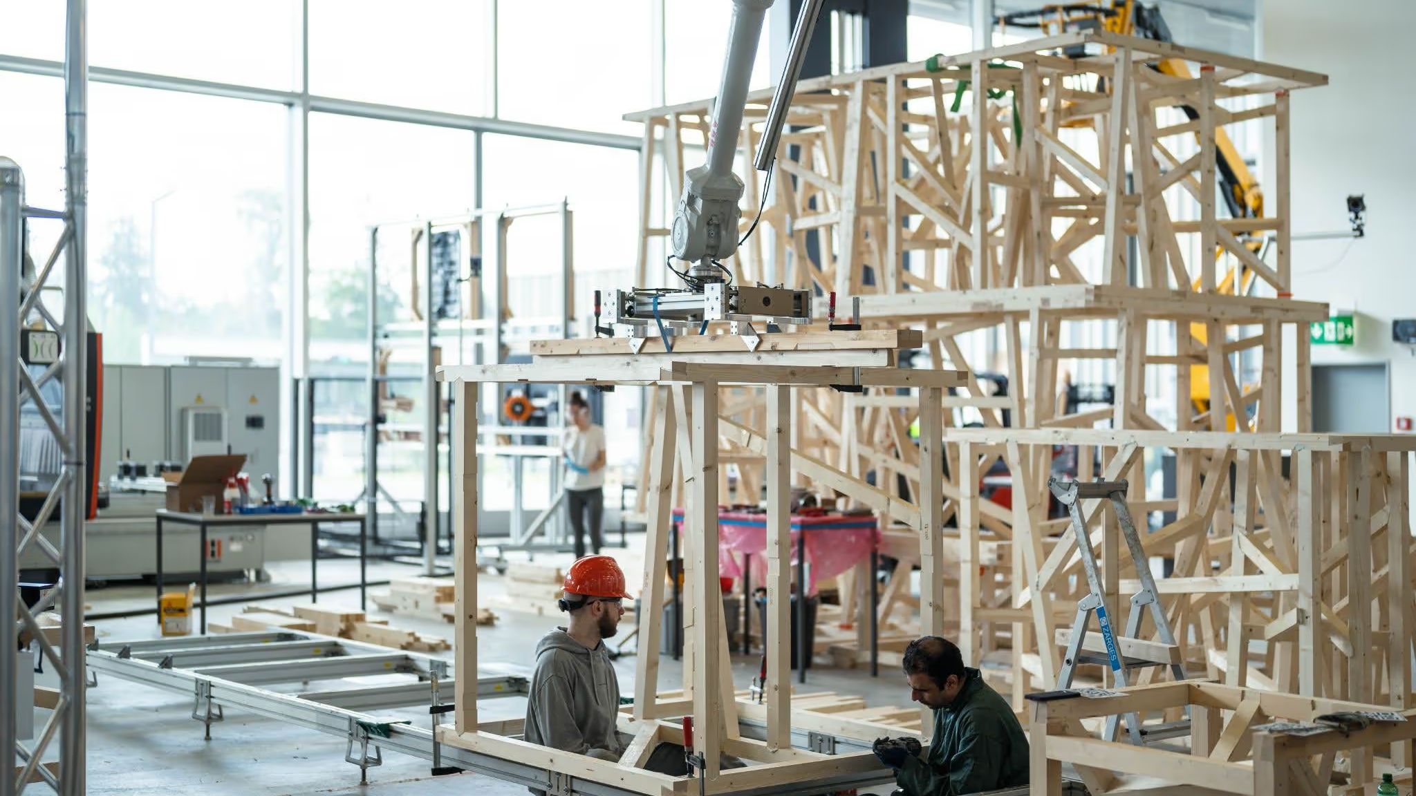 Two researchers working beneath a robot arm that holds a timber frame module in place, with finished modules stacked behind them in a laboratory