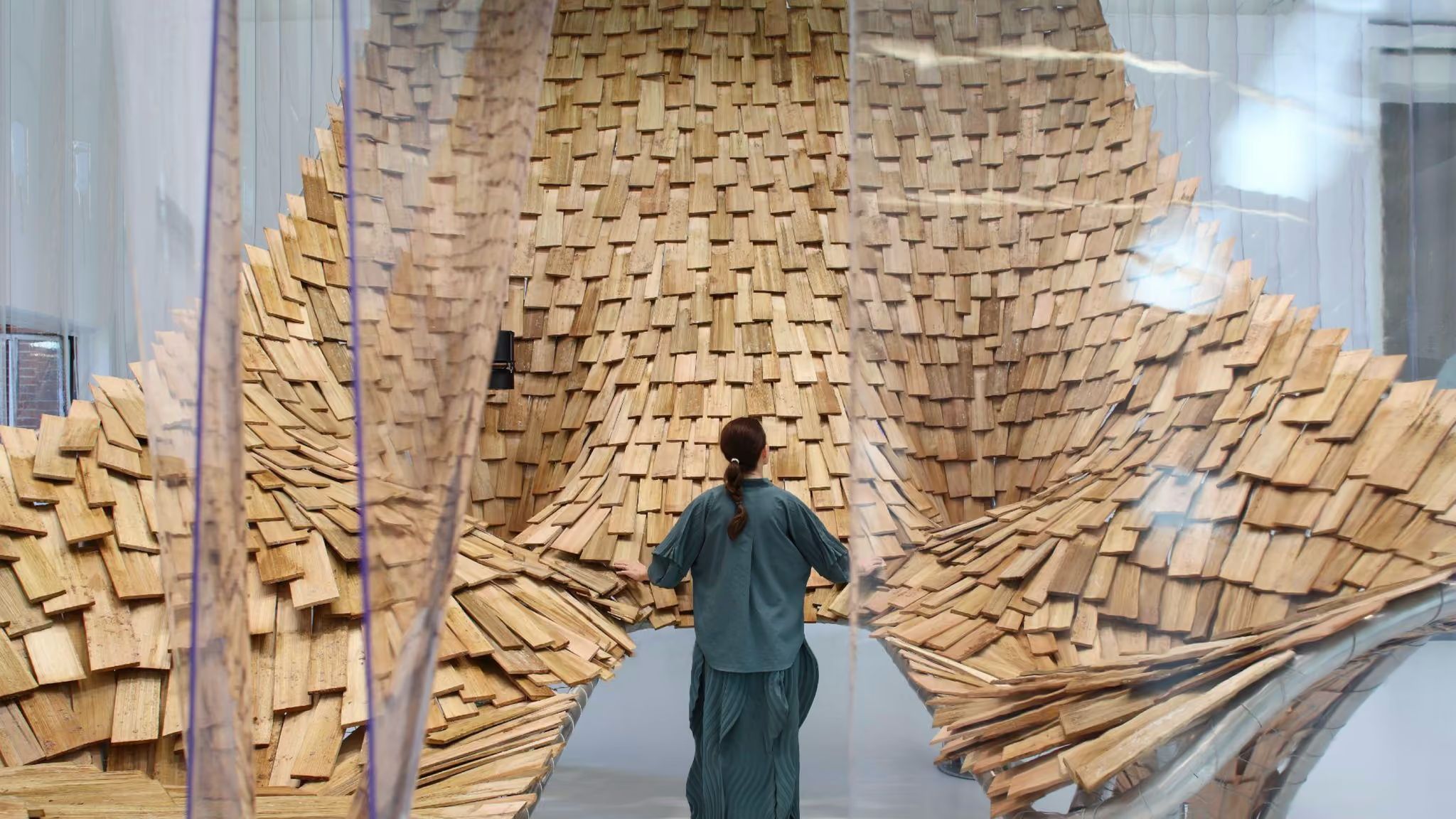 A visitor stands inside a sweeping, curved landscape of overlapping wooden shingles, seen through translucent hanging curtains