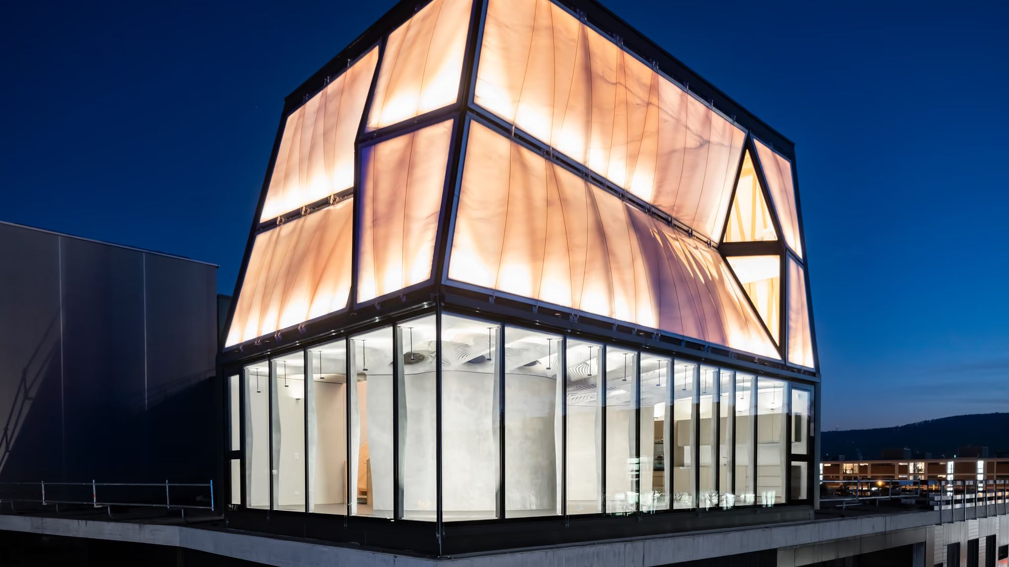 The upper floors of DFAB HOUSE at night, a faceted translucent facade glowing above a glazed storey