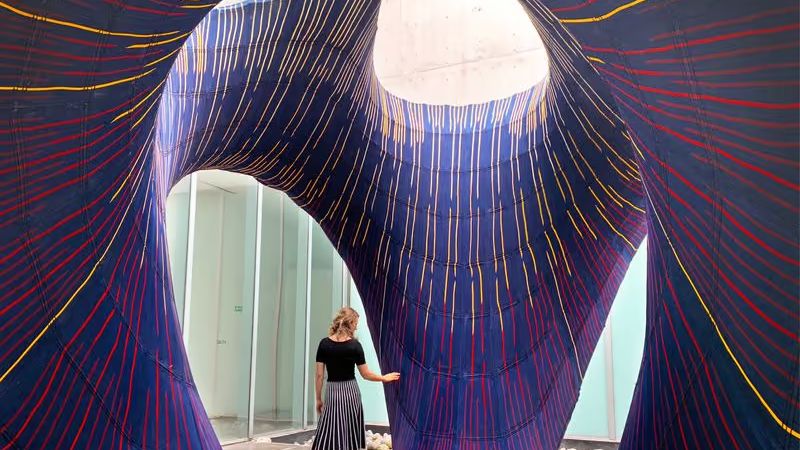 A woman standing inside a sweeping concrete shell lined with a purple knitted textile