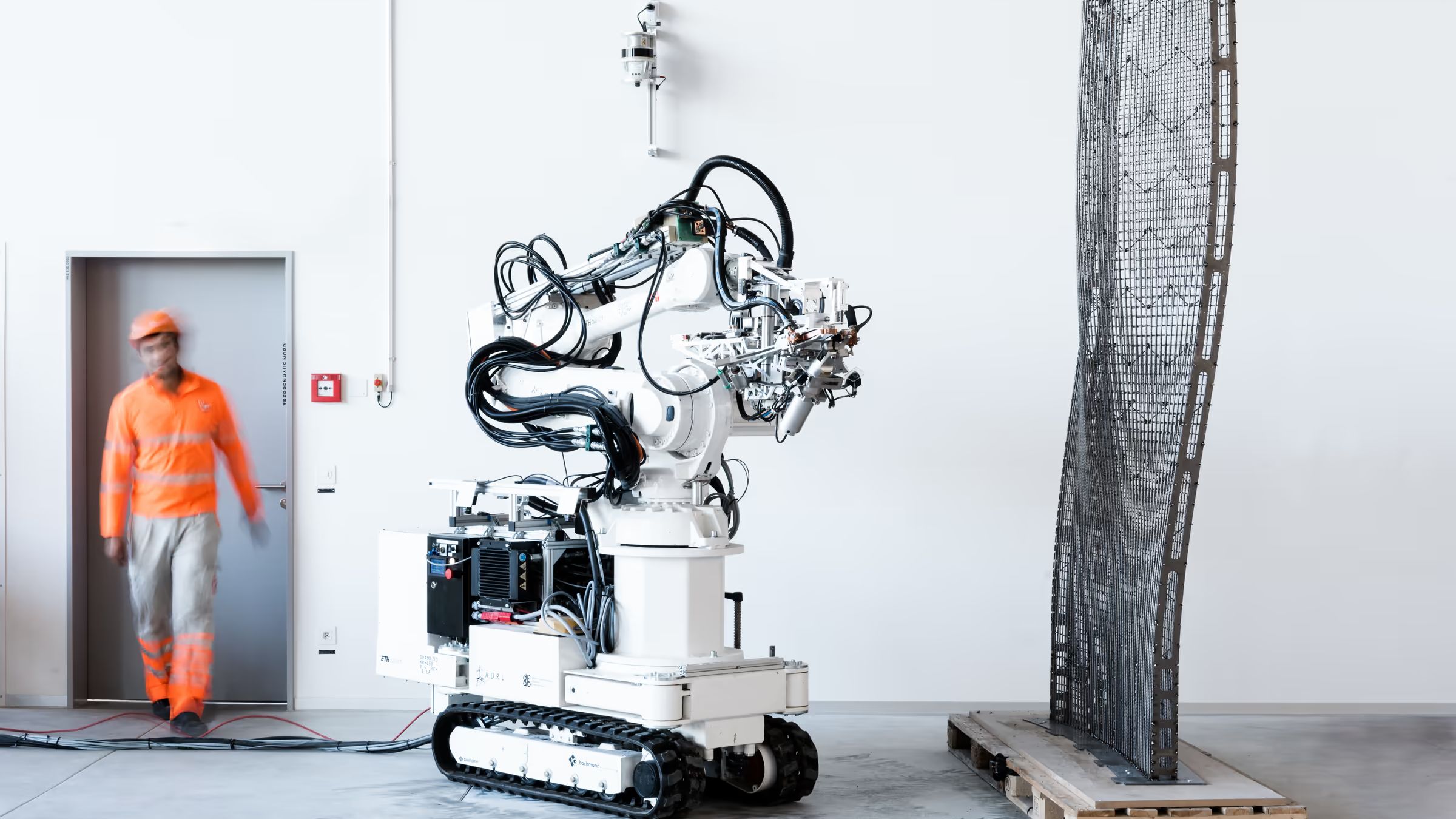 A worker walking past a tracked robot that stands beside a tall, twisting steel mesh it has fabricated