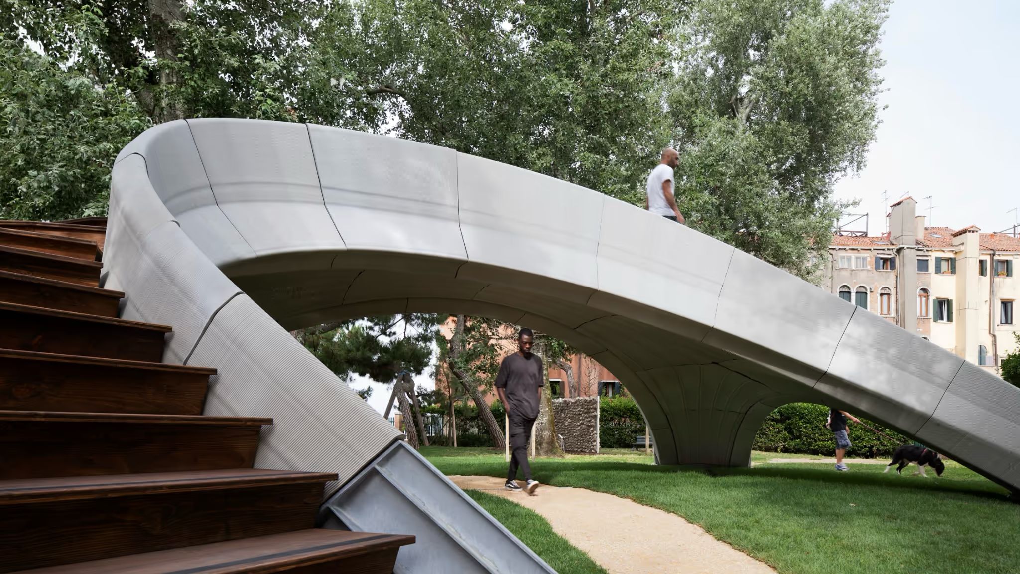 A grey arched concrete footbridge with timber steps in a tree-lined garden, people walking beneath it
