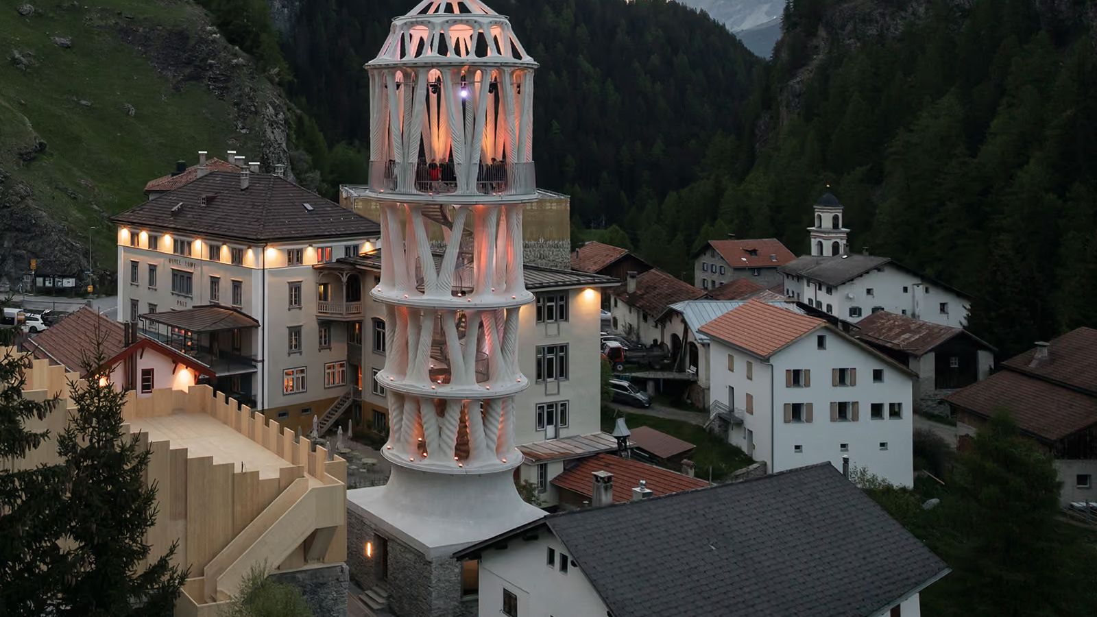 A tall white tower of branching printed concrete columns, lit from within at dusk, rising between houses in a mountain village