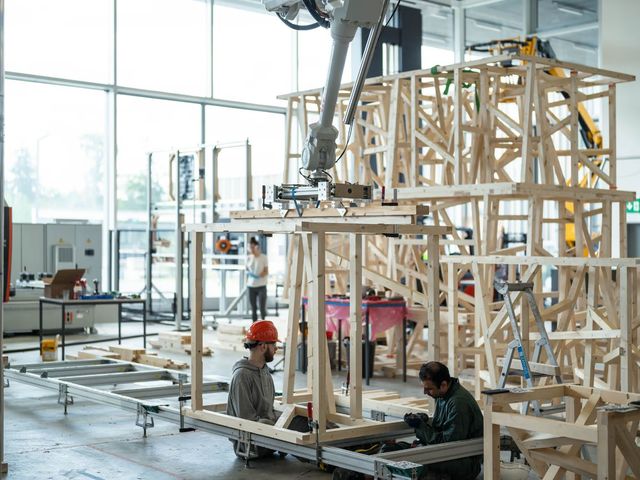Two researchers working beneath a robot arm that holds a timber frame module in place, with finished modules stacked behind them in a laboratory