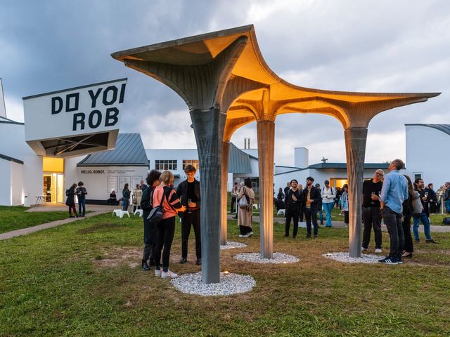Visitors at dusk around a pavilion of slender concrete columns and thin slabs lit warm from below, in front of the Vitra Design Museum