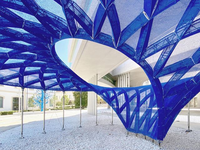 A blue knitted ribbed shell with a large oculus, raised on slender steel props outside a museum